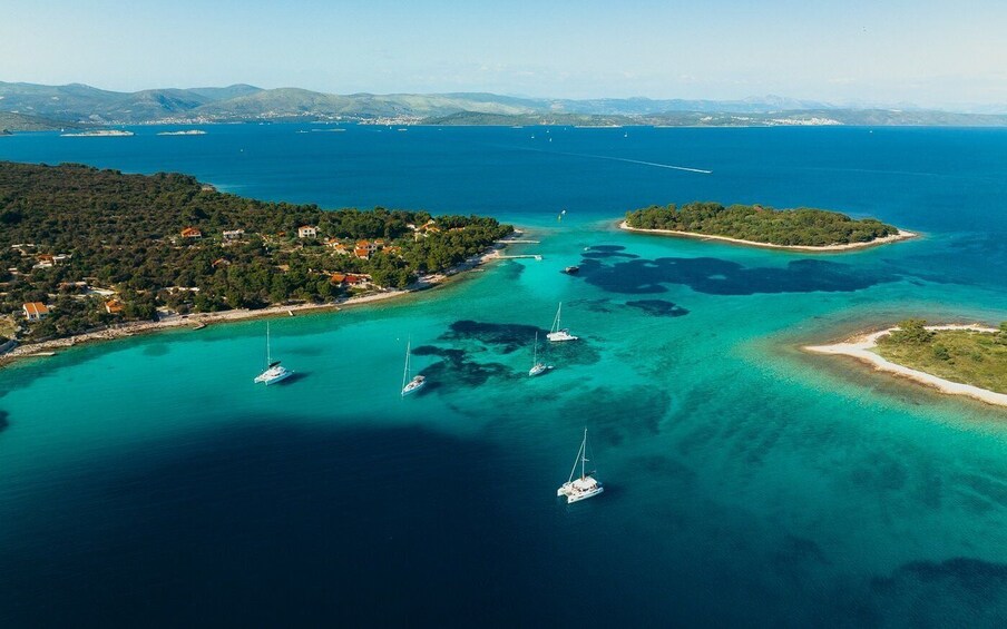 An aerial view of a coastal area with clear turquoise waters, several sailboats, and a small island with dense vegetation. 1