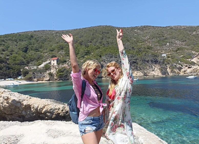 Two people standing by a rocky shoreline with clear blue water and a hilly landscape in the background. 5