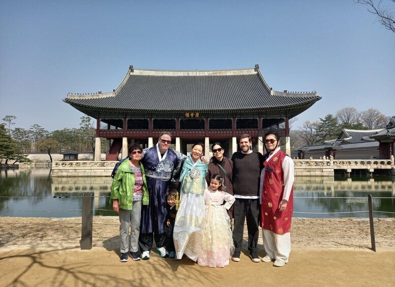 A group of people in traditional attire standing in front of a historical building with a tiled roof and a pond in the background. 2