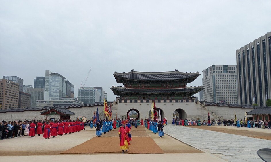 A traditional Korean ceremony with participants in red and blue attire, a historic gate, and modern buildings in the background. 1