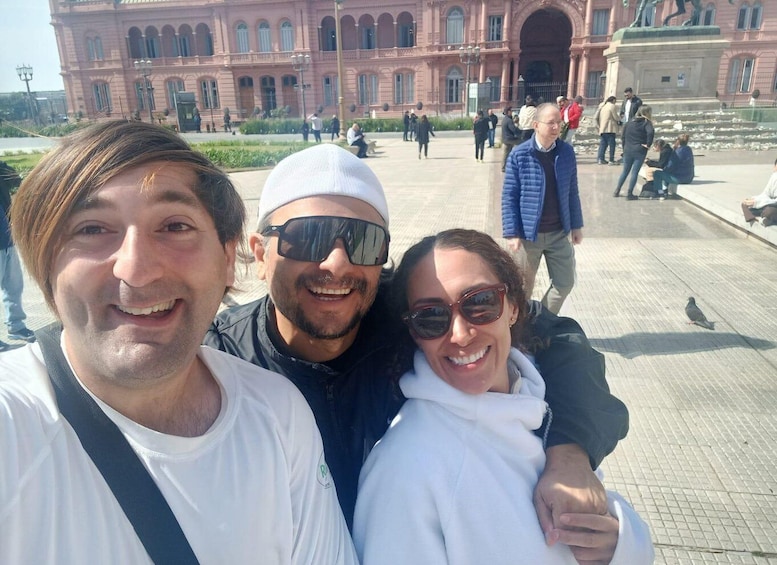 Three people taking a selfie in front of a large pink building with arched windows and a statue in the background. 3