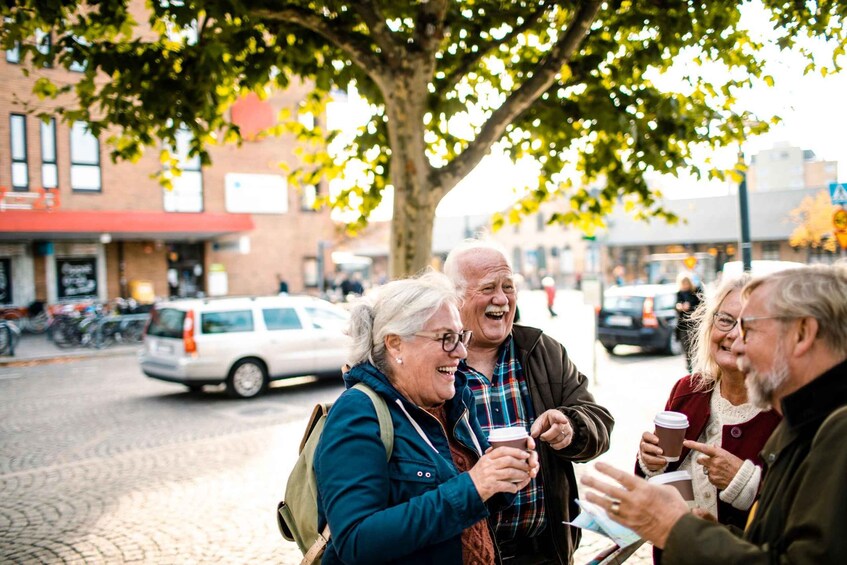 A group of people standing on a cobblestone street, holding coffee cups, with a tree and buildings in the background. 1