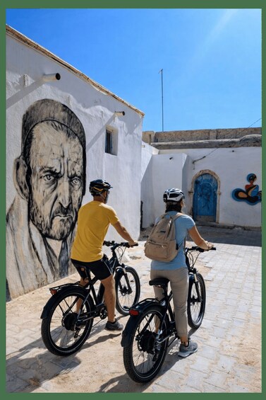Two people on bicycles in front of a building with a large mural and a blue door. 2