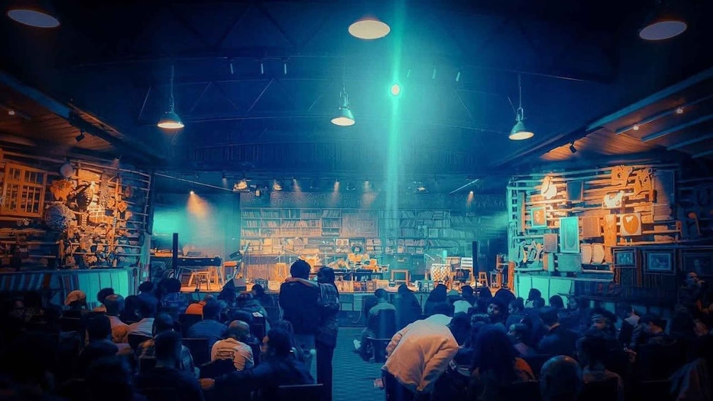 A crowded indoor venue with people seated and standing, illuminated by multiple hanging lights. 3