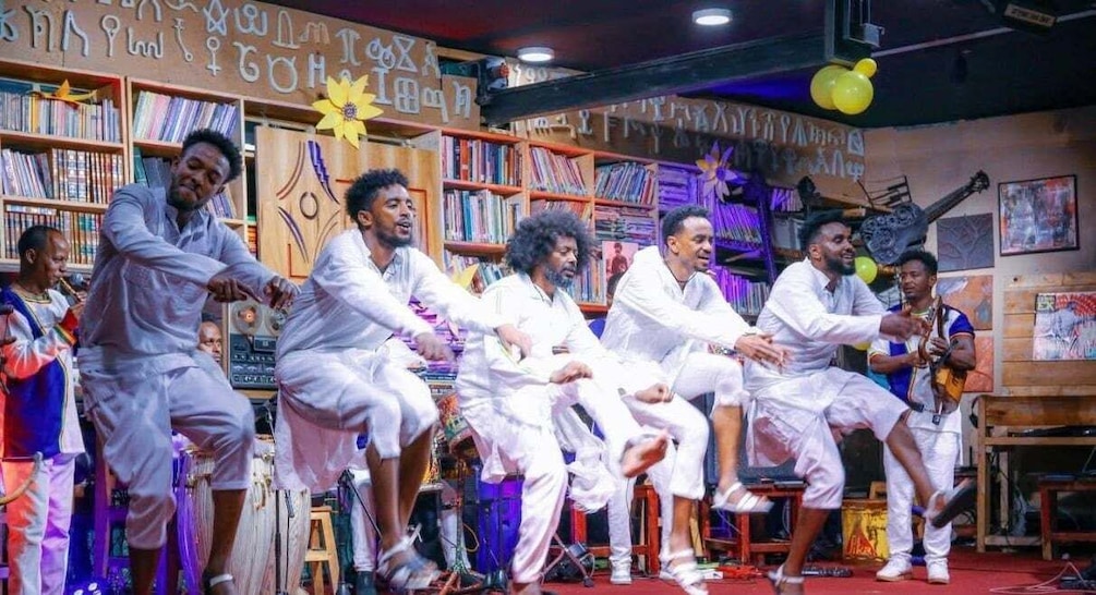 A group of men in white traditional attire performing a dance in a room with bookshelves and musical instruments. 1