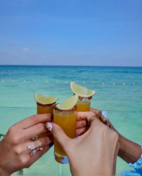 A person holding up three glasses of a yellow beverage with lime slices and a rim of red seasoning, against a backdrop of a clear blue sea and sky. 5