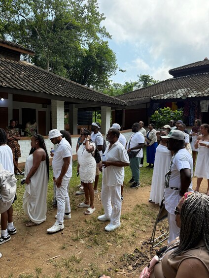 A group of people in white attire gathered outdoors, standing on a dirt ground with buildings in the background. 5