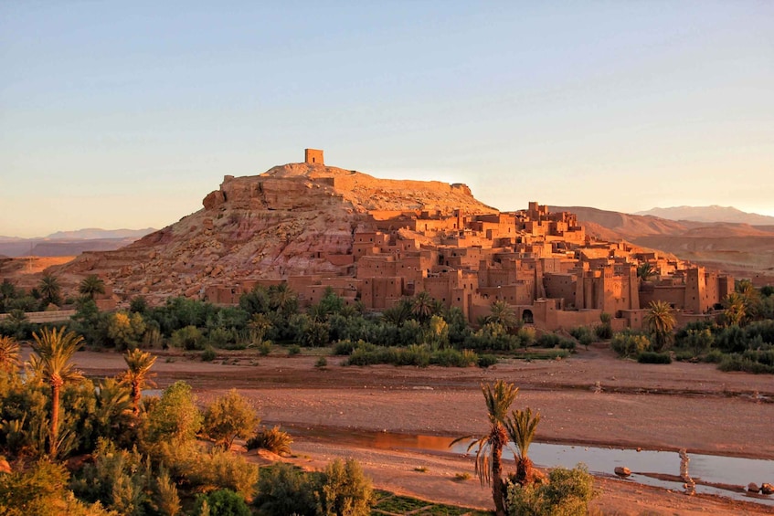 A desert landscape with a fortified village on a hill, surrounded by sparse vegetation and a riverbed. 1