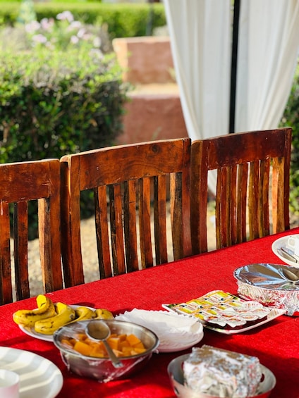 A table with a red tablecloth, wooden chairs, and various food items including bananas, cheese, and a covered dish. 5