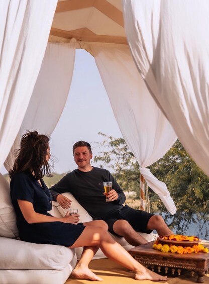 Two people sitting on a couch under a canopy, with a drink and flowers on a table. 1