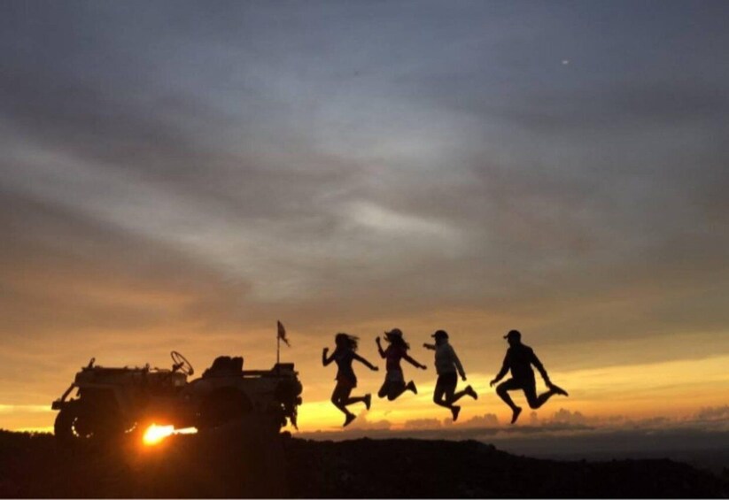 A sunset with silhouettes of people jumping and a vehicle in the foreground. 1