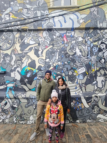 En familie poserer foran en detaljeret mural med forskellige figurer og scener. 4