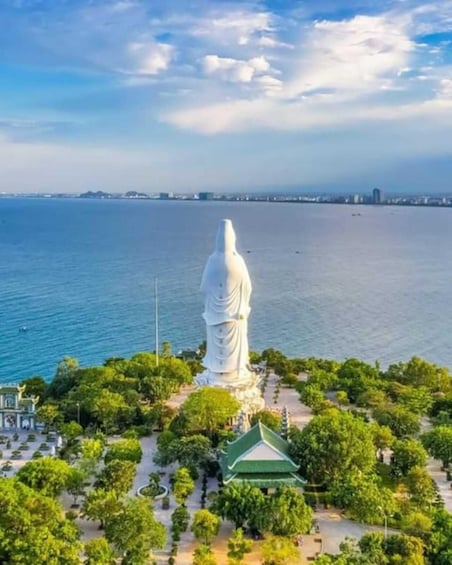 A large white statue on a hill overlooking a body of water, surrounded by greenery and a traditional building. 3
