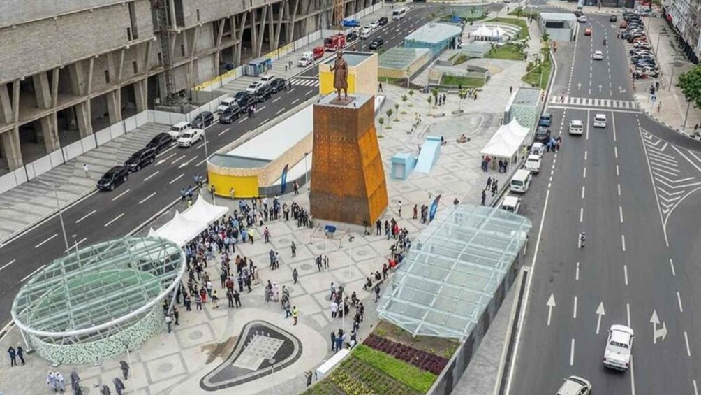 A busy urban square with a prominent monument, a glass structure, and a busy road with vehicles. 5