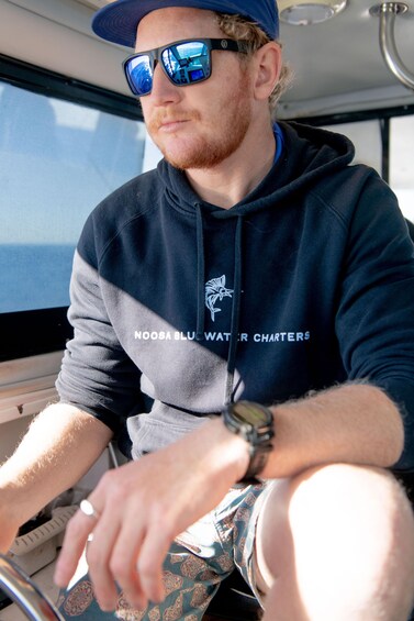 A person wearing a hoodie with 'NOOSA BLUE WATER CHARTERS' text, driving a boat. 5