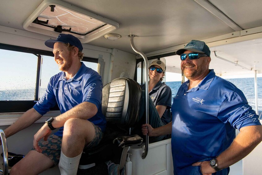 Three people on a boat, wearing blue shirts with logos, and a steering wheel in front. 2