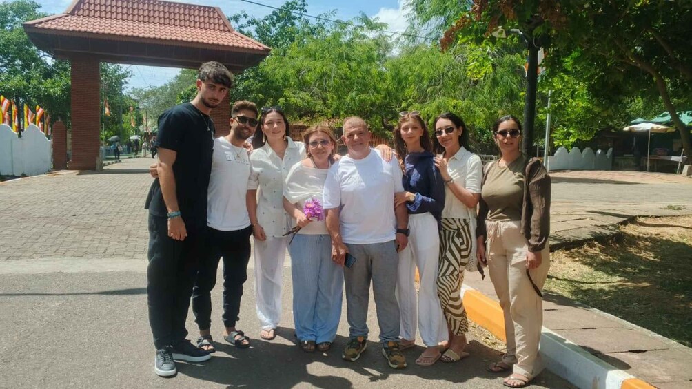 A group of people standing in a public space with a tiled roof structure and greenery in the background. 2
