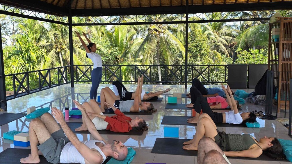 A group of people practicing yoga on mats in an outdoor setting with a thatched roof and palm trees in the background. 1