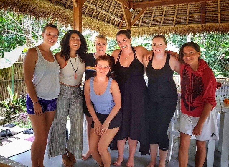 A group of people standing under a thatched roof structure with greenery in the background. 2