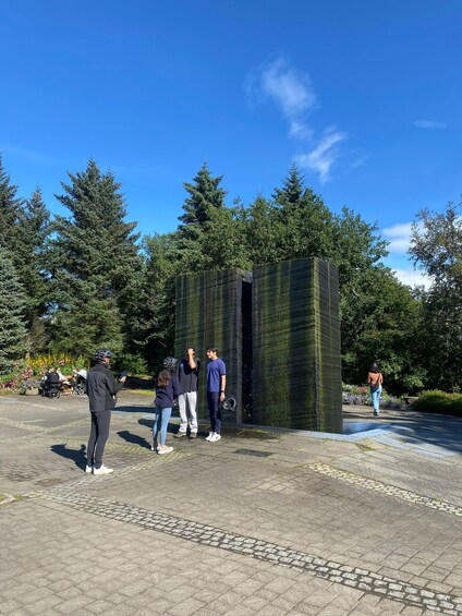 A group of people standing around a moss-covered structure in a park with trees and flowers in the background. 2