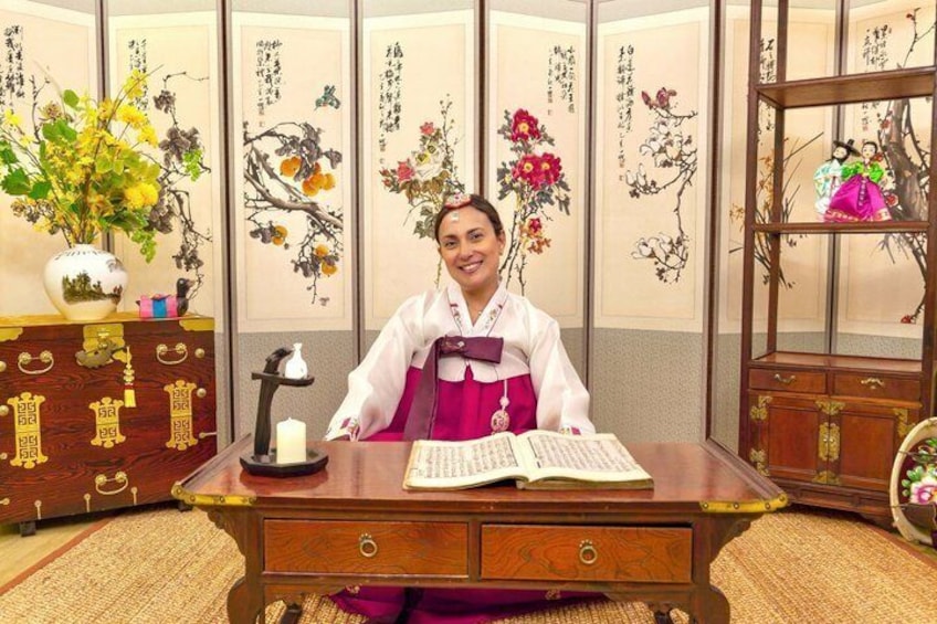 A person seated at a wooden desk with a book, surrounded by traditional East Asian calligraphy and floral artwork. 5