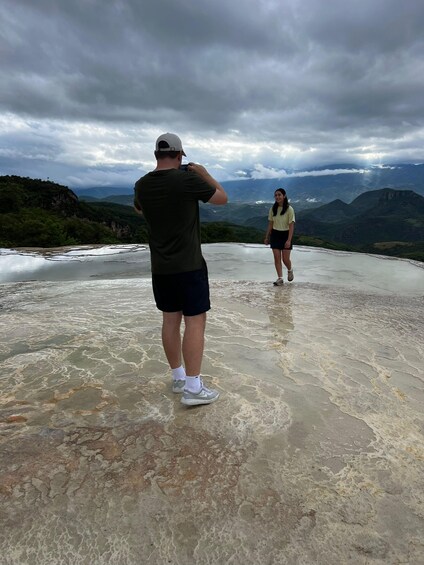 A person taking a photo of another person standing on a rocky surface with a mountainous landscape in the background. 5