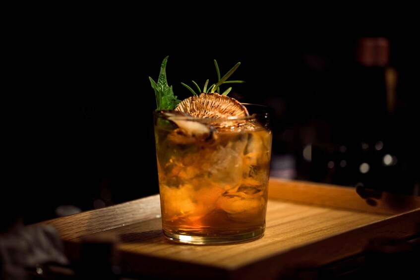 A glass of amber-colored drink with ice cubes and a sprig of rosemary on a wooden bar counter. 1