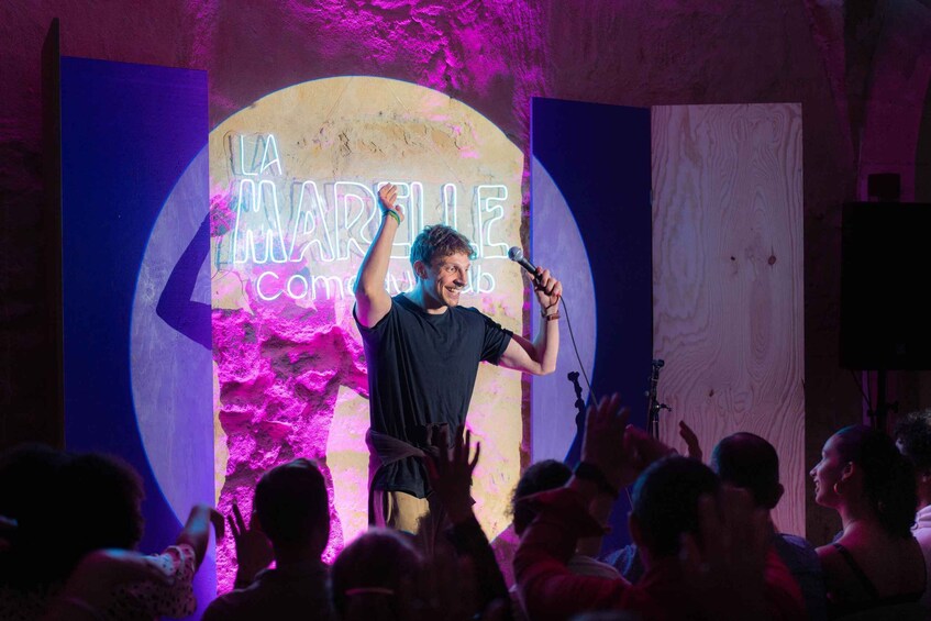 A person on stage with a microphone, a crowd in the foreground, and a neon sign in the background. 2