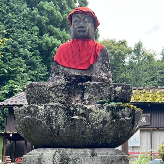 A stone Buddha statue with a red cloth, sitting on a stone pedestal with a bowl in front. 3