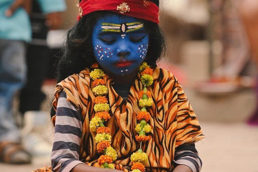 A person with blue face paint, wearing a tiger-striped garment and a red headband, adorned with a garland of orange and yellow flowers. 4
