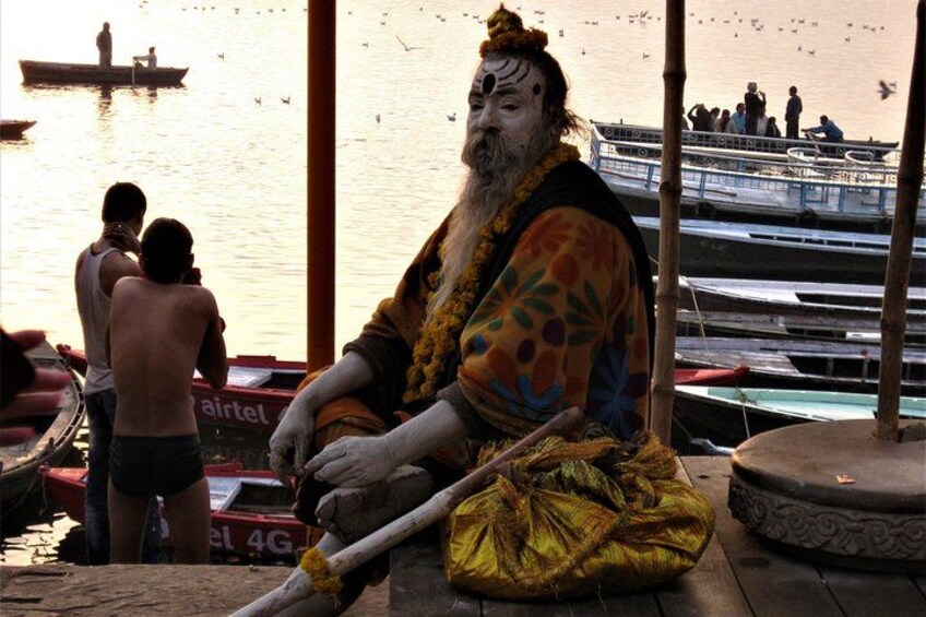 A person in traditional attire sitting by a river with boats and people in the background. 2