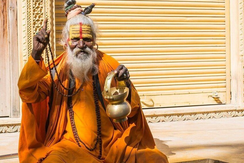 A person in traditional orange attire holding a golden vessel and a string of beads, seated in front of a building with ornate details. 5