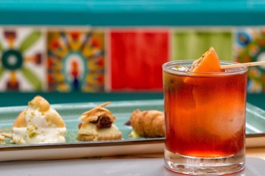Un vaso de bebida roja con una rodaja de naranja, un plato de postres variados y un mural colorido de fondo. 5