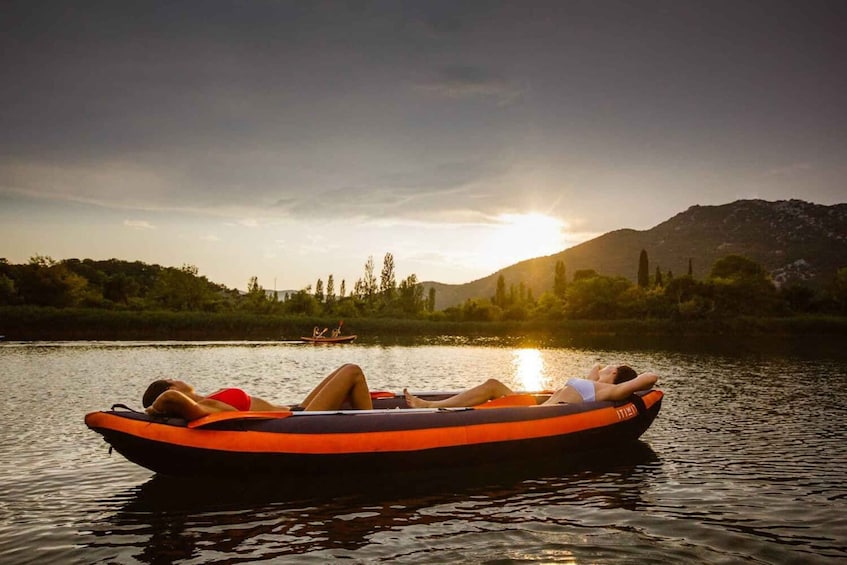 Two people relaxing on a black and orange inflatable boat on a calm river at sunset. 1