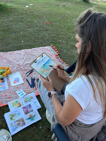 A person painting on a clipboard outdoors with a floral blanket and art supplies. 5