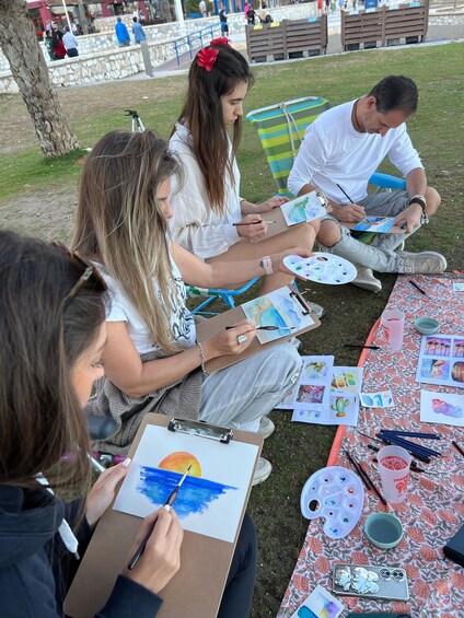 A group of people painting outdoors on a grassy area with a table full of art supplies. 1