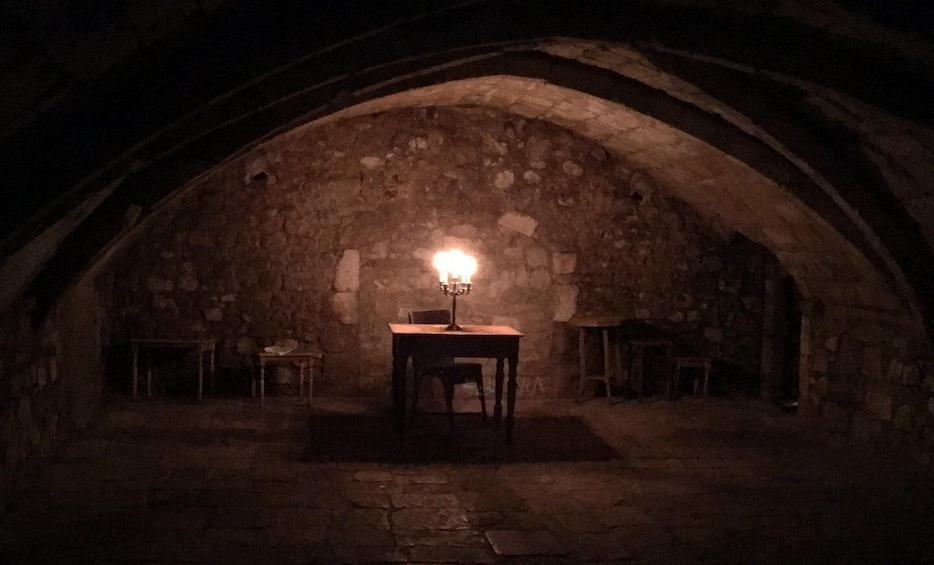 An arched stone room with a table and chairs, a candelabra, and a stone wall. 2