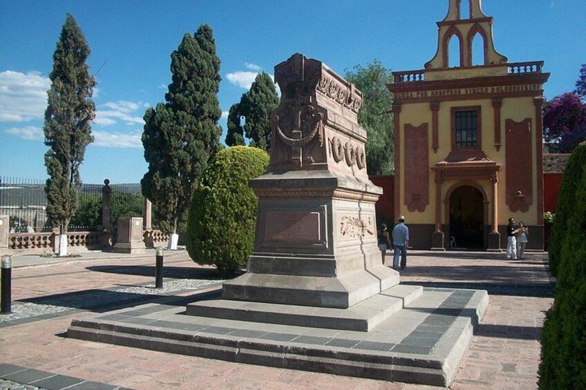 A plaza with a central monument, a bell tower, and trimmed hedges. 3