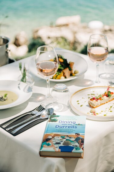 A table set with plates of food, glasses of wine, and a book titled "Dining with the Durrells". 2