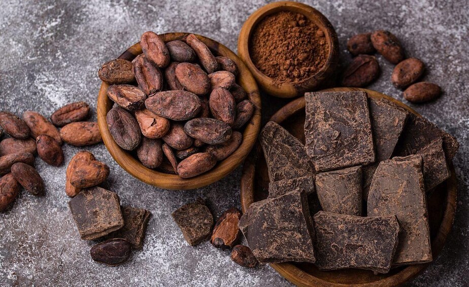 A bowl of cocoa beans, a bowl of cocoa powder, and pieces of chocolate on a textured surface. 3