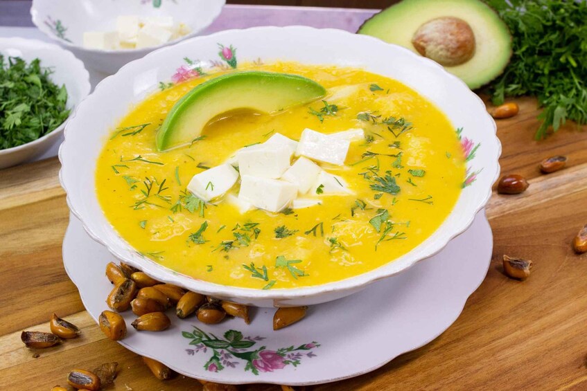 A bowl of soup garnished with avocado slices and herbs, surrounded by ingredients like nuts and herbs on a wooden table. 5
