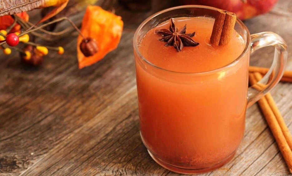 A glass mug of orange-colored beverage with a cinnamon stick and star anise garnish, placed on a wooden surface with autumn leaves and cinnamon sticks in the background. 4