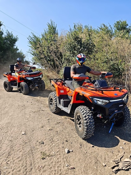 Two people riding orange off-road vehicles on a dirt path with bushes in the background. 2