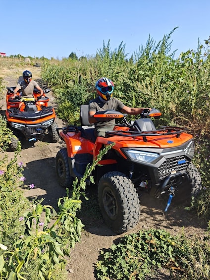 Two people riding orange ATVs on a dirt path surrounded by vegetation. 1