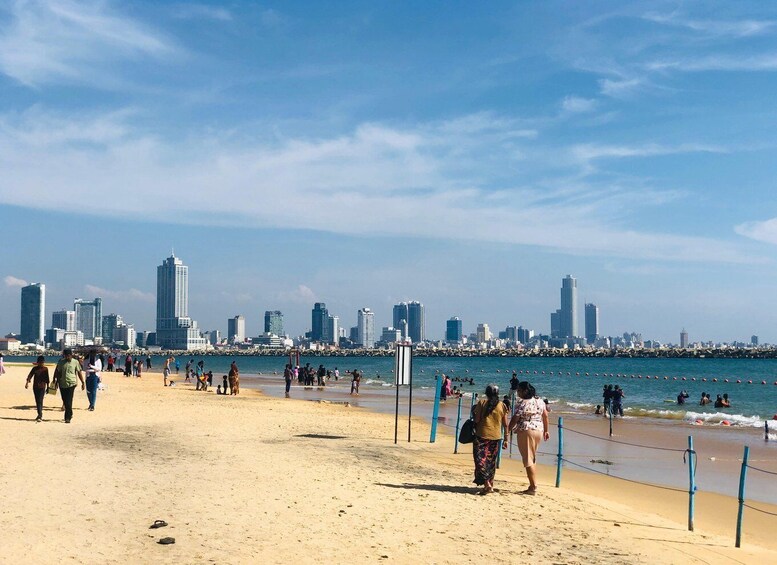 A beach with people walking and swimming, a city skyline in the background, and a pier extending into the water. 2
