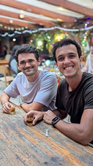 Two men sitting at an outdoor table with drinks, under a covered area with string lights. 3