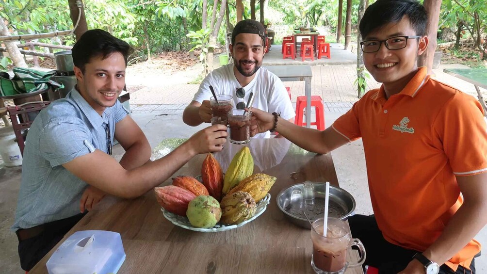 Tres personas brindando con bebidas en una mesa al aire libre, con un plato de frutas y un bol de metal. 1