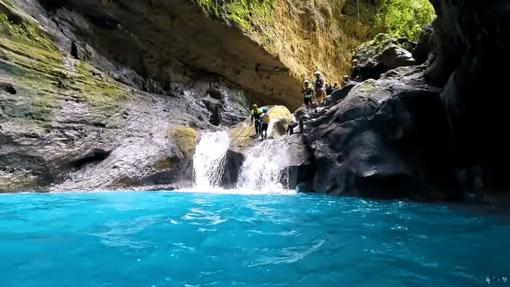 A group of people standing on a rocky ledge above a clear blue pool, with a waterfall cascading down the rocks. 2