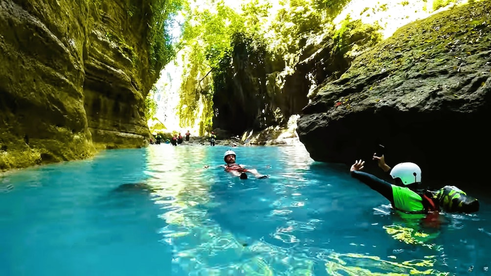 A person floating in a clear blue water body surrounded by rocky cliffs and dense greenery. 5
