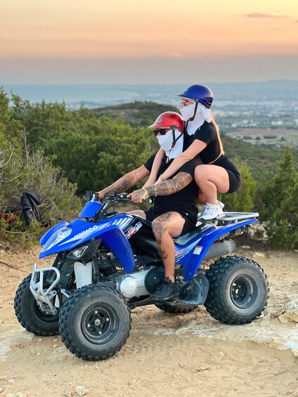 A person on a blue ATV with a passenger, both wearing helmets, in a natural landscape with trees and a distant view of a town. 2